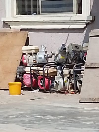 In Olomoda, Nigeria, a collection of portable water pumps is arranged outdoors against a light purple wall, partially obscured by a large piece of plywood. A bright yellow bucket sits on the concrete ground in the foreground to the left. The pumps are stacked in two visible rows, with metal frames supporting them. The upper row consists of at least three pumps, each with a silver engine and a light-colored tank, and some red components. The lower row features more pumps with similar engine and tank designs, predominantly showing red and pink color accents. Above the pumps, a section of a building is visible, featuring a white-trimmed window with dark blinds and a decorative white ledge. The overall scene suggests a storage area or an outdoor marketplace in Olomoda.