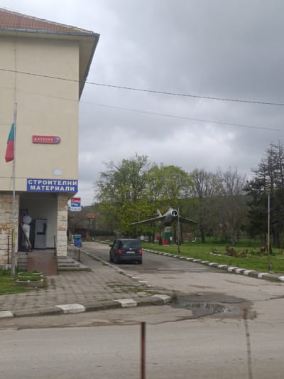 The image depicts a street scene in Komarevo, Bulgaria. A beige building with a red-tiled roof and signs in Bulgarian is on the left, one sign reads "СТРОИТЕЛНИ МАТЕРИАЛИ" which translates to “Building materials”. A Bulgarian flag hangs from the building. A dark grey Volkswagen car is driving on the road toward the center of the image. A decommissioned silver jet fighter plane is mounted on a pedestal in the middle ground, surrounded by grass and trees. The sky is overcast. The road is asphalt, with white and black painted curbs. A large puddle is visible in the road in the foreground.