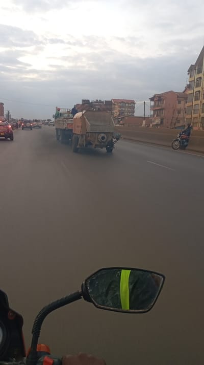 A view from a motorcycle, focusing on the road ahead. A large truck emitting dark smoke is central, followed by other vehicles in the distance. Buildings line the side of the road. A motorcyclist is on the side road. A side mirror with a reflective stripe is visible in the foreground. The sky is overcast.