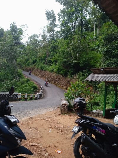 The scene is a dirt path in Cisewu, Indonesia, located at latitude -7.411 and longitude 107.542. The path curves to the left and leads to a paved road winding through dense foliage. Two motorcycles are parked on the dirt path, one facing the road and one facing away. A third motorcycle is parked farther down the path, facing the road. A fourth motorcycle is on the road, heading up the curve. A fifth motorcycle is further up the road, going in the same direction. A person is riding the fourth motorcycle and a person is riding the fifth motorcycle.  Behind the parked motorcycles is a wooden structure with a corrugated metal roof and a green painted support beam. There is greenery and foliage on the left and right sides of the road.