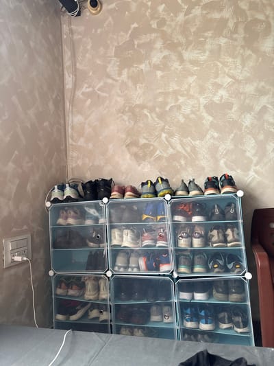 A multi-tiered plastic shoe organizer filled with athletic shoes stands against a textured, beige wall. The organizer is comprised of multiple cubbies, each housing one or two pairs of shoes. The shoes are predominantly sneakers in various colors and styles, including white, black, gray, blue, and red. Some shoes are placed neatly, while others are slightly askew. Above the shoe organizer, a black pipe and white wires are visible against the wall, along with a small, gold surveillance camera mounted in the corner. To the left of the organizer, a white light switch and electrical outlet are present on the wall. A portion of a dark red chair is visible on the right side of the frame. The floor is covered by a gray fabric. The overall scene appears to be an indoor space, possibly a room in a home, located in Chichoga, India.