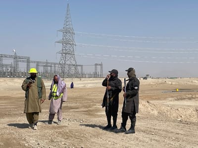The image depicts an open, arid construction or maintenance site for electrical infrastructure in Pir Dust, Afghanistan. Four adult males are present in the foreground and mid-ground.
On the left, two men walk rightward on uneven, dusty brown terrain. The leftmost man wears a light brown traditional outfit, a dark green tactical vest, and a yellow hard hat, looking at a smartphone held in both hands. To his right, a second man wears a light purple traditional outfit, a neon yellow reflective safety vest draped over his shoulders, and a light-colored head covering; he looks downward.
To the right, two armed men stand facing left. Both wear dark uniforms, dark boots, dark baseball caps, and dark face coverings. Each holds a rifle, likely an AK-variant, vertically with the butt on the ground. The man on the far right looks towards the left, while the man closer to the center has his left hand near his ear, possibly operating a communication device.
In the mid-ground, a large electrical substation and multiple tall metal lattice transmission towers dominate the landscape, carrying numerous power lines across the clear, pale blue sky. A single, dark-clad figure is visible walking in the distance near the substation structures. The ground is dry, barren, and uneven, with scattered rocks and small mounds. Distant, hazy mountains are visible on the horizon. On the far right, yellow markers are placed on the ground, and faint structures and a vehicle are discernible in the distance.