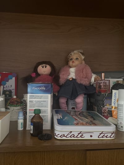 The image depicts a shelf in a wooden cabinet, possibly a dresser or a closet, in Strakonice, Czechia. On the shelf, two dolls are displayed. The doll on the left is a handmade fabric doll with dark brown yarn hair, rosy cheeks, and a pink fuzzy dress. The doll on the right is a more traditional doll with blonde hair styled in a braided bun, blue eyes, and is wearing a pink faux fur boa, a white top, a denim skirt, and pink leggings.
In front of the dolls, there are various items. To the left, a white and blue box labeled "microlife" is visible, which appears to be a medical device or packaging. Next to it is a small brown glass bottle with a green cap, containing a dark liquid. A black coiled hair tie rests on the shelf near the bottle. To the right of the dolls, a pink glass perfume bottle with a gold cap is prominent, labeled "NUXE". Further to the right are several other items, including what seems to be a metal tin with an image of a cityscape and the words "chocolate tuti" printed on it. There are also tubes and bottles of toiletries or medications, one of which is labeled "INDULONA". The overall environment appears to be a domestic setting, likely a bedroom or dressing area.