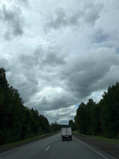 The image presents an overcast sky filled with gray clouds, alongside a road bordered by dense, green trees. A white van travels on the road. The location is not specified as being in the city of Abdes-Urdes, Russia.