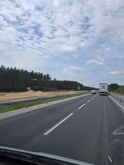The image depicts a highway in Mała Nieszawka, Poland, characterized by a dark asphalt road with white lane markings. On the right side of the road, a silver truck with "Everest" branding is visible. Further down the road, a white car is also traveling. The left side of the highway is bordered by a guardrail and a grassy embankment, beyond which lies a construction site with piles of sand and construction equipment, adjacent to a dense forest of pine trees. The sky is predominantly cloudy with patches of blue peeking through. The perspective suggests the photo was taken from within a vehicle.