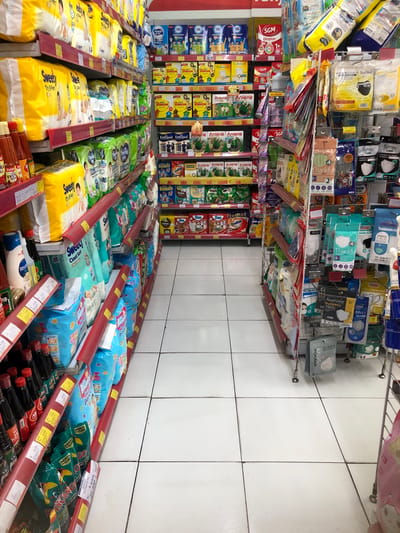 The image shows a retail aisle in a convenience store, likely in Putat, Indonesia, judging by the Indonesian product labels and store signage. The aisle is lined with shelves stocked with various consumer goods. On the left, shelves display bottles of sauces and condiments, including what appears to be soy sauce and chili sauce. Above these, there are packages of baby diapers and formula. The right side of the aisle features shelves with hygiene products, prominently displaying an assortment of face masks in various brands and packaging. Other items visible include baby wipes, and possibly some snacks or processed foods. The floor is tiled with white squares. The lighting appears to be fluorescent, typical of an indoor retail environment. There are no people visible in the image.