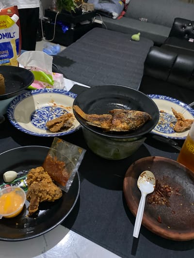 A close-up, eye-level shot shows a dimly lit room in Cibinong, Indonesia, with food laid out on a black tablecloth. The scene is cluttered, suggesting a casual gathering. In the foreground, a black plate holds fried chicken, a small cup of orange sauce, and a sachet of red chili sauce. To the right, a traditional Indonesian mortar and pestle (ulekan) contains leftover chili paste, with a white plastic spoon resting on it. Further back, a blue and white patterned bowl contains a piece of fried chicken and remnants of food. A large black bowl holds a whole fried fish. Behind this, a bottle of iced tea is partially visible. The background reveals a dark sofa, a black leather armchair, and various household items, indicating a residential setting.