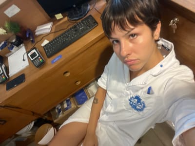 A person in a white shirt, possibly an employee, looks at the camera near a wooden desk. The desk has a keyboard, two payment terminals, a smartphone, a mouse, and some office supplies. A roll of bubble wrap, cardboard box, and white bowl are behind the desk. Keys are to the left of the person. A tattoo is visible on their arm. The image is slightly rotated.