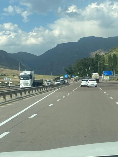 The image depicts a highway scene near Kushminor, Uzbekistan, under a partly cloudy sky. In the foreground, a paved road with white lane markings stretches towards distant mountains. Several vehicles are visible on the road, including a white semi-truck with a trailer in the left lane, and a line of cars, primarily white sedans, in the right lanes. To the left of the road, a guardrail separates it from a grassy embankment. In the background, a range of dark, rugged mountains forms the backdrop, with green trees sparsely dotting the lower slopes. A blue highway sign indicating distances to Namangan, Fargona, and Andijon is visible on the right side of the road. The overall impression is a typical daytime view of traffic on a major road in a mountainous region.