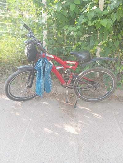 A red bicycle is leaning against a chain-link fence overgrown with green foliage. The bicycle has a black seat, handlebars with grips, and two blue bags with a geometric pattern hanging from the handlebars. The wheels are black with silver spokes. The bicycle is parked on a dirt and asphalt surface. Sunlight casts shadows on the ground, suggesting it is daytime. The scene appears to be outdoors, possibly in a park or along a roadside in Nurabad, Uzbekistan. No people are visible. There is no visible text in the image.