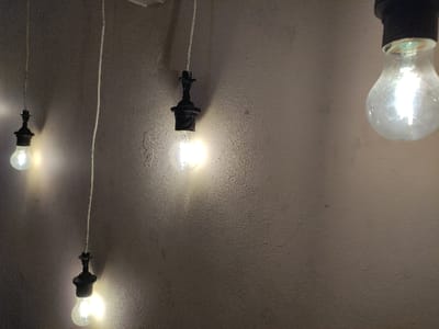 The image displays four illuminated incandescent light bulbs against a textured, light brown wall, characteristic of an interior setting. Three bulbs are fully visible, each suspended by a white electrical wire from an unseen overhead point. The leftmost bulb hangs approximately two-thirds down the frame. To its right, a second bulb is positioned higher, roughly in the middle of the frame. A third bulb is visible in the bottom-center of the image, suspended lower than the second. A fourth, partially visible bulb, also illuminated, is cropped in the upper right corner. All bulbs emit a bright, warm light, casting a glow on the adjacent wall surface. No individuals, additional subjects, or specific actions are present. The precise location within Staufenberg, Germany, cannot be determined from the visual information provided in the image.