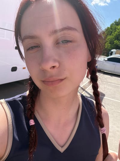 A close-up, slightly angled selfie of a fair-skinned person with reddish-brown hair styled in pigtail braids. The hair is secured with pink hair ties. The person wears a dark blue top with a tan trim. The background shows a parking lot with white vehicles, possibly a bus or van, visible and green trees above. The location is unknown and not confirmed to be in Bekhterevo, Russia.