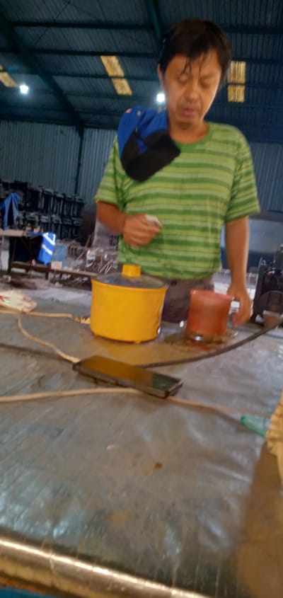 A man in a striped green and white shirt stands next to a table with a yellow cooking pot, a red container, and an object connected to a power cord. The table surface is gray and possibly metallic. The man has a blue and black bag slung over his shoulder and is holding a white object in his hand. The location appears to be indoors, possibly a warehouse or industrial space, as suggested by the corrugated metal walls and exposed lighting. Other metal objects are visible in the background, but are too far away to be distinguished. The city and country are Sumput, Indonesia.