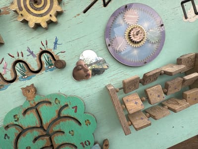 The image is a close-up, overhead shot of a light green, weathered wooden surface with various interactive toy elements attached. The surface appears to be a wall or a large panel, possibly part of a children's play area.
Prominently featured are several wooden toys designed to engage children. In the upper left quadrant, a large, multi-layered wooden gear with a yellow, spiraling maze inside is mounted. Below this, a black, wavy track with small red and blue painted dragonflies and cattails is embedded in the wood, with a small wooden bead running along the track.
To the right of the maze gear, a circular transparent disc with intricate gear designs is affixed. A small, cloud-shaped mirror is placed below this, reflecting a portion of a young child's face and the surrounding environment, which appears to be outdoors with trees and possibly a playground structure.
In the lower half of the image, a large, green, wooden clock face shaped like a tree is visible. The clock face has cut-out numbers (5, 7, 8, 9, 10) and several carved tree branches. A small wooden owl figure sits atop the tree.
To the right of the clock, a chain of wooden blocks is arranged diagonally. These blocks are square and made of natural wood, each featuring a small engraved image such as a rocket, a hot air balloon, a ship, or a star pattern. These blocks appear to be threaded onto a metal rod, allowing them to slide.
The overall scene depicts a well-loved and worn interactive play surface, likely designed for sensory and motor skill development. The location is stated to be in Zaliznychne, Ukraine, although this detail cannot be verified from the image itself. The lighting suggests it is daytime, possibly outdoors due to the reflection in the mirror. There are no people directly visible, only the reflection of a child.