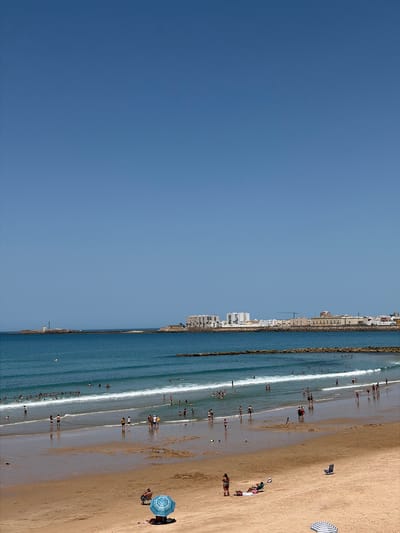 This is a photograph of a beach scene in Cádiz, Spain. The image depicts a wide expanse of sandy beach with numerous people enjoying the water and sun. The ocean is calm with gentle waves lapping the shore. Several individuals are wading or swimming in the shallow water, while others are walking along the beach. 

On the sandy portion of the beach, there are beach umbrellas and towels, indicating a typical day at the seaside. Some people are relaxing under umbrellas, while others are sunbathing or playing. The cityscape of Cádiz is visible in the background, with buildings lining the coast, suggesting an urban beach setting. The sky is clear and blue, indicating a sunny day, likely during daylight hours. The overall atmosphere appears relaxed and leisurely, characteristic of a vacation or a day out at the beach. There is no visible text in the image.