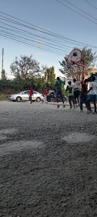 The scene depicts an outdoor basketball activity in Frere Town ward, Kenya, under a clear, light blue sky. Approximately eight individuals, appearing to be male adolescents or young adults, are present on an unpaved dirt and gravel court. A central figure, dressed in a green shirt and dark pants, is airborne directly beneath a red-framed basketball hoop with a white backboard and net, indicating a jump during active play. Other individuals are positioned around the hoop, observing or participating. In the left background, a silver sedan is parked, partially obscuring a darker vehicle. Dense green trees and foliage form a natural backdrop along the left and center. Numerous overhead utility lines are visible against the sky, originating from a utility pole on the far left.