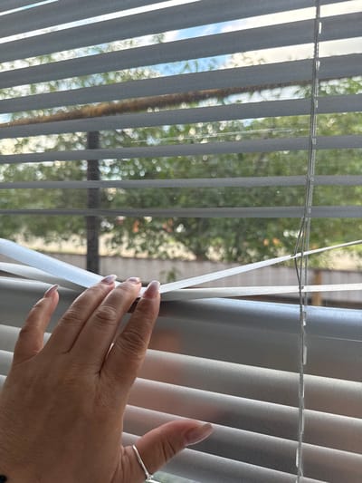 In this media file, a person's hand is seen reaching towards a set of gray vertical blinds. The hand, adorned with a silver ring on the index finger and light-colored fingernail polish, is positioned to the right of the frame. The blinds are partially open, allowing a view of the outdoors. Through the gaps in the blinds, greenery and foliage are visible, suggesting an outdoor setting with trees. The lighting indicates it is daytime, likely with sunlight filtering through the leaves and causing a bokeh effect in the background. The scene appears to be captured indoors, looking out through a window with blinds. The blinds themselves are made of a matte material and are organized in a way that suggests they are adjustable. The focus is on the hand interacting with the blinds, with the background softly blurred. There is no visible text in the image. The image does not provide specific cues to pinpoint the exact location beyond the general context of looking out a window at greenery, but has been tagged with Novokubansk, Russia.