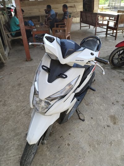 This image depicts a casual outdoor gathering space with a concrete floor in Meunasahpantonlabu, Indonesia. In the foreground, a white Honda scooter is parked, facing the viewer. The scooter exhibits dust and dirt, with the "HONDA" logo visible on its black seat and "Combi Brake" text on the right handlebar housing. A portion of a red motorcycle's front wheel and fender is visible to the right of the white scooter.

In the midground, to the left, a group of four men are present around a wooden table with chairs. One man in a teal shirt is seated at the table, looking downwards. Another man is partially obscured behind the table, also seated. A third man in a dark, possibly striped, shirt is seated further left, facing right. A fourth man in a blue shirt is seated further back and to the right of the table, engaged in drinking or eating from an object held to his mouth. Objects on the table include a blue plastic bag and a small white item, possibly a cigarette pack or phone.

The background features a wooden structure forming the rear of the covered area. To the far right, an empty wooden table with two matching benches is visible. Beyond the covered area, an unpaved outdoor environment with green foliage and distant structures is apparent under natural light.