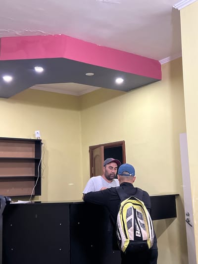 The image depicts an indoor scene, possibly a reception or service area, with two individuals interacting. The room has yellow walls and a dark grey ceiling with pink accents. There is a black counter in the foreground.
One individual, wearing a grey t-shirt and a baseball cap, is standing behind the counter, appearing to be speaking with the other person. The second individual, wearing a black jacket and a blue baseball cap, is facing away from the camera, with a yellow and black backpack on. The backpack has visible branding, including "Bab".
To the left of the counter is a tall, dark shelving unit with empty shelves. On top of the shelving unit, a small white device with a wire is visible. A door is partially visible on the right side of the image.
The overall impression is that of a transaction or conversation taking place in a functional, utilitarian space. The location in Kadamjay, Kyrgyzstan, is not visually evident from the image itself.