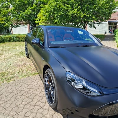A matte black sports car is parked on a paved area next to a grassy lawn. The car's sleek design is evident, with sharp lines and a low profile. The front headlight assembly is visible, featuring intricate LED lighting. The car's wheel is a prominent feature, showcasing a multi-spoke design with orange brake calipers peeking through. The tire has "MICHELIN PILOT SPORT S S" printed on its sidewall. The interior of the car, visible through the tinted windows, appears to be a reddish-brown leather.
In the background, a large, lush green tree with dense foliage provides a natural contrast to the car. Beyond the tree, a building with a light-colored facade and windowed entrance is partially visible. A person wearing a pink shirt and blue shorts can be seen through the entrance, interacting with someone inside. The paved ground is made of interlocking concrete blocks arranged in a honeycomb pattern. The scene suggests a sunny day, likely in the late morning or early afternoon, given the bright lighting and clear shadows. The overall setting appears to be a suburban or public area, possibly a parking lot or the grounds of a business or residential complex in Rastatt, Germany.