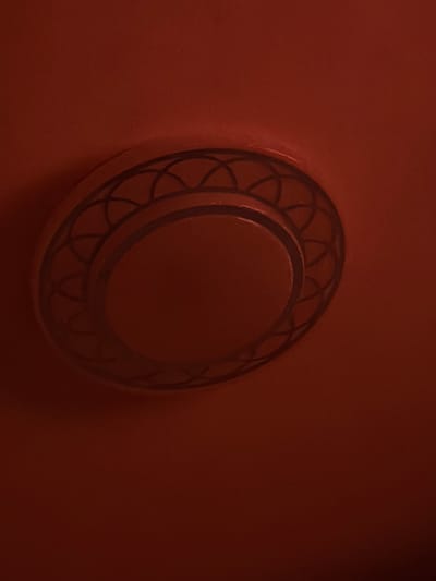 An interior scene located in Rain, Germany, depicts a circular decorative fixture mounted on a flat, uniformly colored surface. The entire visible environment, including the fixture and its background, is rendered in a deep reddish-brown hue, resembling terracotta or rust. The fixture, positioned slightly off-center in the upper portion of the frame, features a central, solid, circular disc that is slightly recessed or raised. This central disc is surrounded by a wider, concentric ring adorned with a repeating, dark-lined geometric pattern of interconnected arcs, forming a lattice-like motif. A thinner, unpatterned ring separates the central disc from this decorative outer ring. The scene is dimly lit, casting subtle shadows and highlights, with a faint illumination visible along the left edge of the fixture. No human figures, objects, or activities are present in the image.