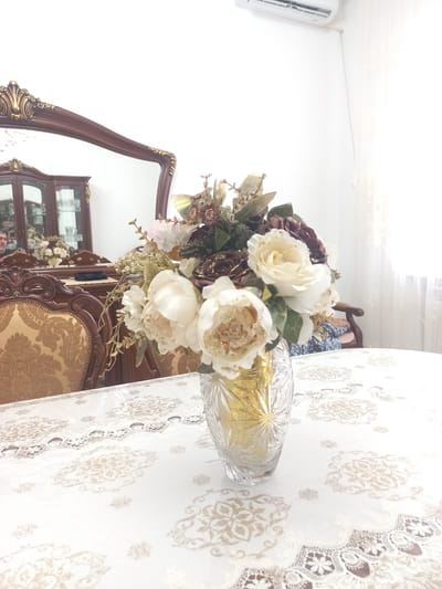 The image depicts an indoor scene, likely a dining room, featuring a table covered with a white tablecloth adorned with intricate gold and cream patterns. On the table, a crystal vase holds a bouquet of artificial flowers, predominantly white and deep burgundy, accented with gold foliage and glitter. The vase is clear crystal with a faceted design, and it contains what appears to be gold decorative elements.
In the background, a large, ornate dark wood mirror with gold trim is visible. Reflected in the mirror is a dark wood china cabinet with glass doors, displaying glassware and other decorative items. A portion of a dark wood dining chair with patterned upholstery is visible in the foreground to the left. Another chair, with a wooden frame and blue and white patterned fabric, is partially visible to the right, behind the vase. An air conditioning unit is mounted high on a white wall in the upper portion of the image. The scene is well-lit, with light appearing to come from a window to the right, which is covered by sheer curtains.
There are no discernible elements in this image that allow for the identification of the specific location within Zyazikov-Yurt, Russia. The scene is a general interior setting.