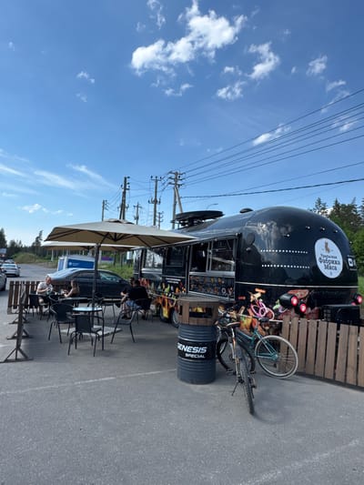 The image depicts an outdoor food truck setup under a bright blue sky with scattered white clouds, likely in Finskiy Briz, Russia. A prominent black, oblong food truck, styled like an Airstream, serves as the central subject. Its side features the white Cyrillic text "Уральская Фабрика Мяса" (Urals Meat Factory) with an animal logo, along with "СТОRЕ" and colorful graphics of various food items including meats, sausages, and vegetables. White star patterns also adorn its exterior.
In front of the food truck, a casual outdoor dining area is arranged with several black metal tables and chairs, shaded by two large, light-colored patio umbrellas. Multiple people are seated at these tables, appearing to be patrons enjoying a meal or break in the open air.
To the right of the food truck, a mountain bike is parked, leaning against a wooden fence or barrier. A black barrel labeled "GENESIS SPECIAL" sits nearby. The ground is a paved asphalt surface, possibly a parking lot or roadside area.
In the background and stretching across the clear sky, numerous power lines are visible, supported by several utility poles. Green trees line the horizon behind the setup. The bright sunlight casts clear shadows, indicating it is daytime, likely mid-morning or afternoon, with pleasant, sunny weather. The scene suggests a relaxed roadside eatery or an outdoor café in a semi-rural setting.