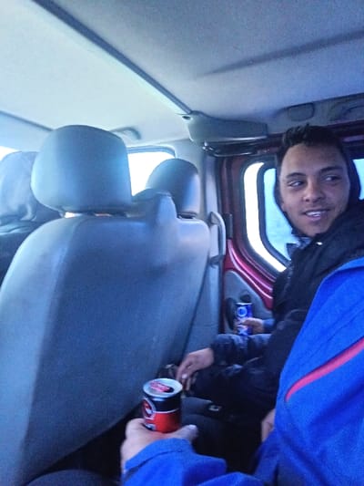 The scene depicts the interior of a multi-passenger vehicle, identified as being in Dzhurovo, Bulgaria. Three individuals are present within the vehicle. In the foreground, a left hand, wearing a blue jacket with a red stripe detail, holds an open red and black aluminum beverage can. To the left, the upper portion of a grey vehicle seat and headrest is visible, partially obscuring an occupant. To the right, a young adult male with dark hair, wearing a dark hooded jacket, is seated. He is looking towards the right side of the frame with a slight smile, and his right hand holds a blue and white aluminum beverage can against his leg. The vehicle's interior features a light-colored ceiling liner, grey synthetic seats, and a red door panel with a window on the right side, through which bright, undifferentiated daylight is visible.
