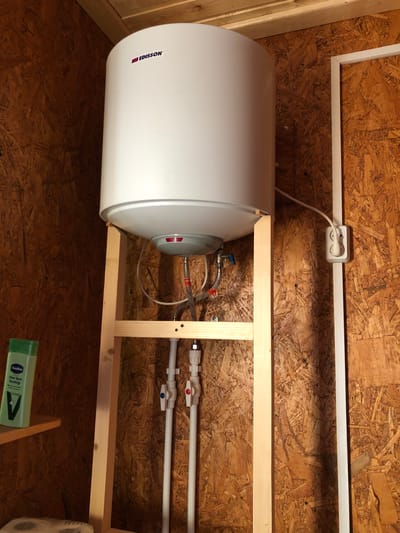 In Uriupino, Russia, an electric water heater, branded "EDISSON," is mounted on a wooden frame against a wall constructed of oriented strand board (OSB). The white cylindrical water heater is positioned above a network of white plastic pipes with red and blue valves, indicating water supply lines. A white electrical cord snakes from the heater to a white electrical outlet mounted vertically on the OSB wall. To the left, a green bottle of Vaseline Aloe Vera lotion sits on a narrow wooden shelf. The scene suggests a utilitarian installation within a storage or utility area.