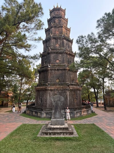 The image depicts the Thien Mu Pagoda in Kim Long Ward, Vietnam, a tall, multi-tiered structure built of dark brick. The pagoda is surrounded by trees. A woman in a light-colored dress stands on a stone structure near the pagoda's base. People are visible further back, near a stone building. The ground is a mix of brick paths and green grass.