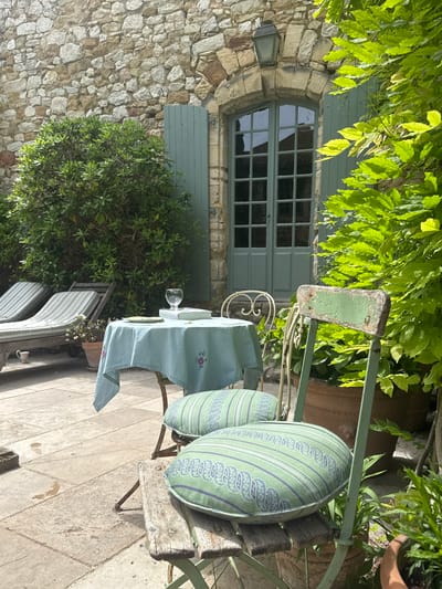 The scene is in an outdoor courtyard in Saint-Victor-la-Coste, France. A stone building with a shuttered window and leafy vines is visible. A small table covered with a light blue cloth and a wine glass is next to a green metal chair with striped pillows. In the foreground, there are sun loungers. The ground is paved with stone tiles.