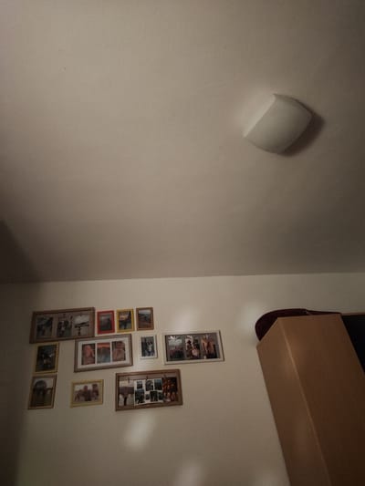 The image shows a wall with several framed photographs displayed. The frames vary in size and color, with some containing multiple smaller photos. The photographs appear to be personal memories, with images of people and landscapes. One frame, located in the upper right portion of the wall, is a light fixture. To the right of the wall is a large wooden cabinet, with a dark object resting on top of it. The overall scene suggests an indoor setting, likely a home or personal space. The lighting appears to be artificial, casting soft shadows on the wall and objects. There are no visible signs of outdoor weather conditions or a specific time of day indicated, beyond the general illumination. No text is discernible in the photographs or on any other elements within the frame. The arrangement of the photos indicates a decorative choice, personalizing the space with captured moments.
