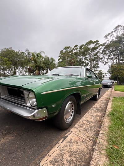 A green classic Ford Falcon car is parked on a road next to a concrete curb. The car is angled toward the camera, showcasing its front passenger side with a round headlight and shiny chrome bumper. Green grass grows on the other side of the curb, with trees filling the background under a cloudy sky. Another darker car is visible further down the road behind the Falcon.