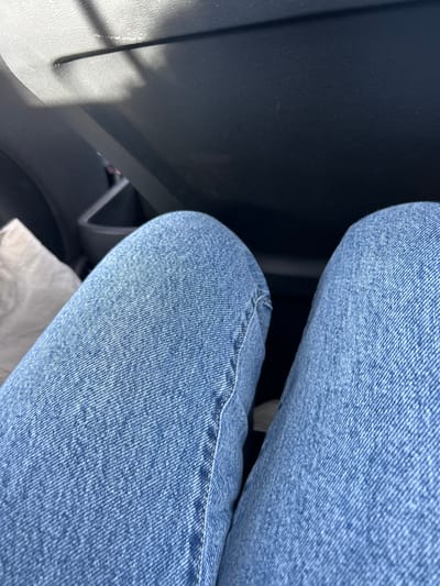 This image depicts an interior view within a vehicle, described as being located in La Boisse, France. The primary subjects are the two legs of an individual, from the mid-thigh down, clad in light blue denim jeans. The legs are positioned parallel, slightly diverging upwards from the bottom of the frame, with the knees prominent. White stitching is visible along the outer seam of the left pant leg. The background consists of dark, textured interior components of a car. The upper section shows a dashboard or glove compartment area, with strong sunlight casting defined rectangular shadows across its surface. A light-colored, crumpled object, possibly a bag, is partially visible on the vehicle's floor to the left of the left leg. No specific actions or interactions are discernible beyond the individual being seated within the vehicle.