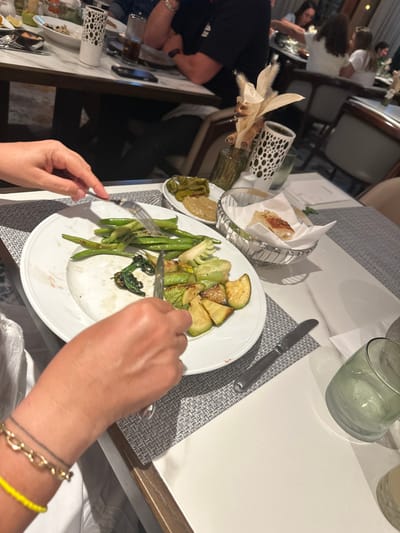 The image captures an indoor dining scene within a restaurant in Daliyat al-Karmel, Israel. The primary focus is a close-up of a person's hands, positioned in the lower-left to mid-frame, actively eating from a white oval plate. The left hand, adorned with a gold-toned chain bracelet and a bright yellow beaded bracelet, holds a fork. The right hand holds a knife, engaged in cutting or preparing a portion of the grilled vegetables on the plate. The plate contains green beans, sliced zucchini, and some dark leafy greens, with visible signs of partial consumption.
The table surface is white, covered by a grey geometric-patterned placemat beneath the main plate. A silver knife rests on the placemat to the right of the plate. Further right, a textured clear drinking glass is visible. Other items on the table include two small white oval dishes holding side items: one contains green pickled vegetables, and the other holds a light brown dip. A metal basket lined with white paper contains pieces of bread. In the mid-ground, a clear glass vase holds dried, light-colored decorative elements, and a tall white cylindrical container with black polka dots is present. The blurred background shows other restaurant patrons seated at separate tables with upholstered chairs, indicating an active dining environment.