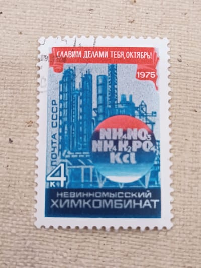 The image is a close-up of a postage stamp. The stamp depicts an industrial scene with tall, blue structures that appear to be smokestacks or factory buildings. In the foreground, a red circle contains chemical formulas: NH₄NO₃, NH₄H₂PO₄, and KCl. There is also a large number '4' in blue with a white outline. Vertical text on the left side of the stamp reads "ПОЧТА СССР" (Pochta SSSR), which means "USSR Post". A red banner across the top of the stamp has white Cyrillic text that reads "СЛАВИМ ДЕЛАМИ ТЕБЯ, ОКТЯБРЬ!" (Slavim delami tebya, Oktyabr'!), meaning "We praise you with deeds, October!". The year "1975" is printed in a red rectangle on the right side of the stamp. The bottom of the stamp has white Cyrillic text on a dark blue background that reads "НЕВИННОМЫССКИЙ ХИМКОМБИНАТ" (Nevinnomysskiy Khimkombinat), meaning "Nevinnomyssky Chemical Combine". The stamp is perforated around the edges and is placed on a textured, beige fabric background. There is no indication of the city of Onaldy, Turkmenistan, in the image or on the stamp itself.