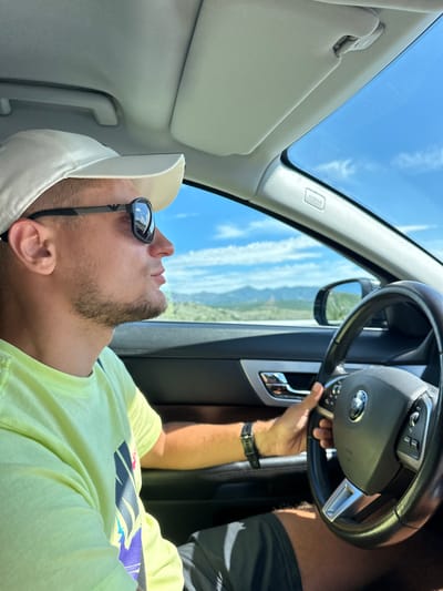 A medium shot captures a young man driving a car on a sunny day, with a blurred mountainous landscape visible through the windshield and side windows. He is wearing a white baseball cap, dark sunglasses, and a light green t-shirt. His right hand is on the steering wheel, and his left arm rests on the car door. The interior of the car shows a black dashboard and leather seats. The background outside the car reveals a clear blue sky with scattered clouds, distant mountains, and vegetation. The specific location is not discernible from the image, and no elements definitively place the scene in Pelev Brijeg, Montenegro.