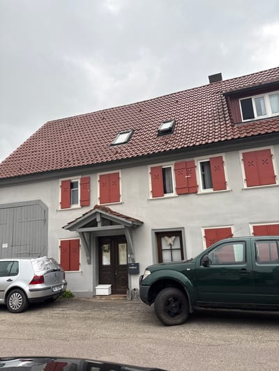 The image captures a cloudy day in Untermünkheim, Germany, focused on the exterior of a two-story house with a red-tiled roof. The house features numerous red shutters on its light grey facade, some of which are open, revealing windows. There are several skylights on the roof, and a chimney is visible. A small covered entrance with a grey frame shelters the main door, which is dark brown. Next to the entrance is a white box.
In front of the house, a silver Volkswagen Golf is parked on the left, its rear window covered with a reflective material. To the right of the Golf, a dark green SUV is parked, partially obscuring the lower part of the house. A black mailbox is mounted on the wall to the right of the entrance. The ground is paved with asphalt. The overall impression is of a residential property under an overcast sky.