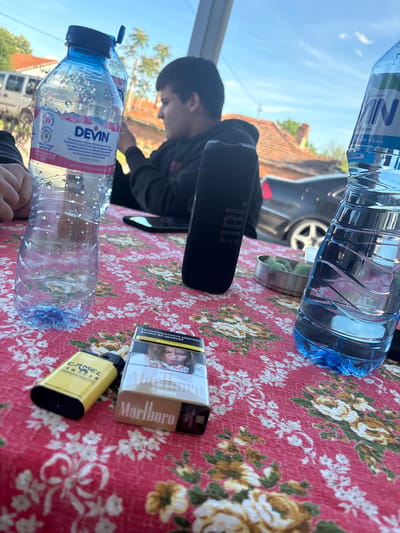 The scene takes place in Blatets, Bulgaria, on a floral-patterned red tablecloth. A young person is seated behind the table, partially visible. The table holds a JBL speaker, two bottles of water (one labeled "DEVIN" and the other "IN"), a pack of Marlboro Gold cigarettes, and a yellow Camel lighter. A car is partially visible in the background.