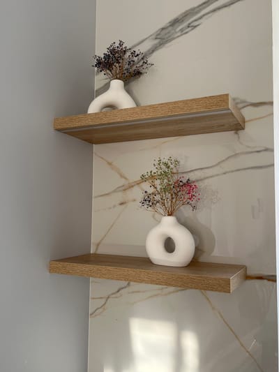 This is a medium shot, eye-level, indoor photograph of two wooden shelves mounted on a wall with marble-like tiling. The wall on the left is painted a solid, light gray. The shelves are approximately one foot apart.
On the top shelf, to the right of center, sits a white, C-shaped vase containing a bouquet of dried, dark purple wildflowers. The vase has a matte finish.
On the bottom shelf, to the right of center, sits a white, torus-shaped vase with a circular opening in the center. This vase also has a matte finish and contains a bouquet of dried wildflowers in various colors, including green, pink, and dark blue. The shadow of this vase and its contents is cast on the tiled wall behind it.
The overall scene suggests a minimalist and decorative interior design. There are no people visible in the image. The location is not specified in the image or its metadata.