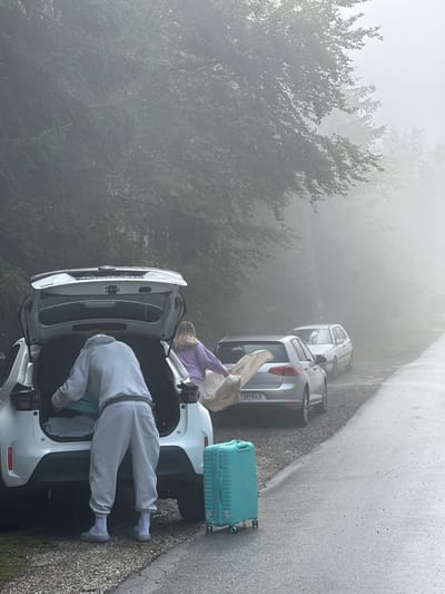 In the misty mountain environment of Trešnjevik, Montenegro, two individuals are observed unloading a white vehicle. A person, wearing a light grey hooded sweatshirt and matching sweatpants, is actively placing items into the open trunk of the car. To the right of this individual, another person, clad in a light purple long-sleeved shirt and white pants, is holding and appears to be folding a light-colored fabric. Beside the car, a bright turquoise suitcase with wheels stands on the wet asphalt road. Two other cars are parked further down the road, partially obscured by the fog. The surrounding landscape is dense with trees, and the air is heavy with mist, creating a subdued and somewhat ethereal atmosphere.