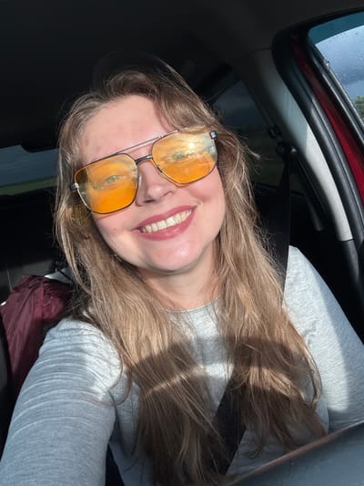 The image shows a blonde woman in a car, wearing sunglasses. The reflection in her glasses reveals a view of Verkhnie Iziverki, Russia. She is wearing a gray shirt and a seatbelt. The window is wet with raindrops.