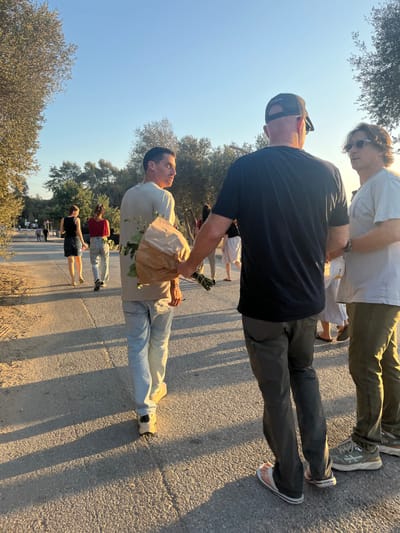 The image depicts an outdoor scene on a paved path during what appears to be late afternoon, characterized by a clear sky and strong, low-angle sunlight casting long shadows. The specific location in Brener Regional Council, Israel, is not discernible from the visual information provided.
In the immediate foreground, three adult males are present. On the left, a male with short dark hair, wearing a light-colored short-sleeved shirt and light blue jeans, walks away from the camera. He holds a bundle of green foliage, partially wrapped in brown paper, in his left arm and turns his head to look back over his right shoulder. He wears light-colored athletic shoes. To his right, a second male is seen from behind, wearing a black short-sleeved shirt, dark grey cargo-style pants, and a dark baseball cap worn backward. He wears white flip-flops. To the right of him, a third male with light brown wavy hair and sunglasses wears a light-colored short-sleeved shirt and olive green pants, with light-colored athletic shoes. He is turned towards the male in the black shirt, appearing to be in an interaction.
Further down the path in the midground, several other individuals are walking away from the camera. These include a female with dark hair wearing a black top and dark skirt, and another female with dark, curly hair wearing a red t-shirt and light blue jeans, who also appears to be carrying foliage. Additional indistinct figures are visible in the background.
The environment consists of a textured, light-colored paved path. On both sides, the path is bordered by mature trees and dense green foliage, suggesting a park or natural area. The sky is clear and blue, and the direct, warm sunlight from the right creates prominent, elongated shadows of the people and vegetation on the path.