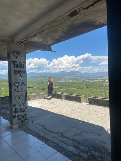 A woman in a black dress stands on a concrete balcony overlooking a valley in Gornji Kokoti, Montenegro. The balcony is part of an unfinished concrete structure, with graffiti visible on a supporting pillar. The woman is seen from behind, walking away from the camera towards the scenic landscape. The background features rolling green hills, mountains, and a bright blue sky with white clouds.