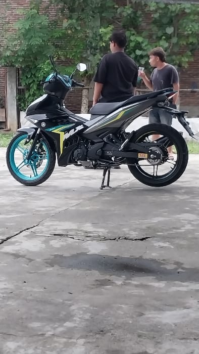 The image depicts an outdoor scene in Mali Guyui, Indonesia. A black Yamaha Exciter motorcycle, featuring green and yellow graphic decals and a distinctive turquoise front wheel rim, is centrally positioned on a cracked and uneven concrete surface. The motorcycle is oriented towards the left and supported by its kickstand.
Two adult males are present. The first male, viewed from the back, wears a black short-sleeved t-shirt and dark lower garments. He stands to the right of the motorcycle, partially obscuring the seat. The second male, seen from his left side, wears a dark grey short-sleeved t-shirt and dark shorts. He holds a clear plastic cup in his right hand, extended forward, and is looking towards the first male, suggesting an interaction or conversation.
The background consists of dense green foliage from trees or shrubs, with portions of a brick wall visible behind the vegetation. The concrete ground displays significant cracking and visible dark, wet patches. The scene is illuminated by ambient daylight.