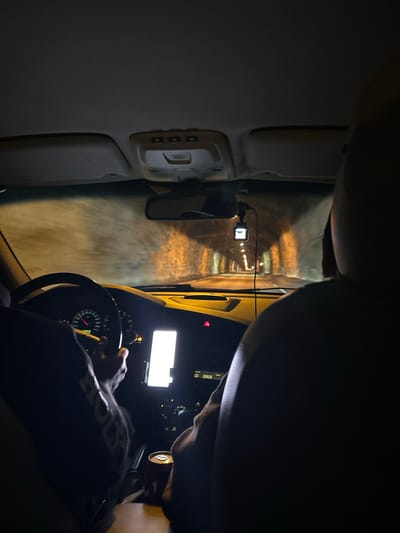 The image shows the interior of a vehicle from a first-person perspective, as if the viewer is the driver or passenger. The scene is dimly lit, suggesting it is nighttime. The car appears to be in motion, possibly traveling through a tunnel given the illuminated walls and overhead lights receding into the distance.

In the foreground, the steering wheel and a portion of the dashboard are visible. A person's hands are on the steering wheel. A screen, possibly a GPS or entertainment system, is illuminated on the dashboard. A small camera, likely a dashcam, is mounted on the windshield, facing forward. The interior of the car includes parts of the ceiling, a rearview mirror, and a sun visor.

The external environment, seen through the windshield, is a tunnel with alternating lights casting a pattern on the walls. The texture of the tunnel walls suggests it might be made of concrete or rock.

The overall impression is one of nighttime travel through an enclosed passage. There is no clear indication of weather conditions, but the lighting suggests artificial illumination. The image captures a common experience of driving at night, potentially in a rural or mountainous area given the tunnel. No explicit emotions are conveyed, but the focus on the driving experience is evident. There is some faint text visible on the person's shirt, appearing to be an acronym or word, but it is largely obscured by shadow and motion blur. The location context of Horda, Norway, would suggest a possibility of driving through mountainous terrain where tunnels are common.