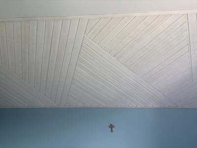 The image shows a section of a white wooden paneled ceiling with a geometric pattern of planks arranged at various angles. The planks are narrow and appear to be tongue-and-groove. Below the ceiling is a blue wall. Centered on the wall, below the paneled ceiling, is a small, brown, metallic cross. The scene is indoors and appears to be residential. The image is a photograph taken from a low angle looking upwards. There is no information about the specific location in the city of Дольче Вита Кп, Russia.