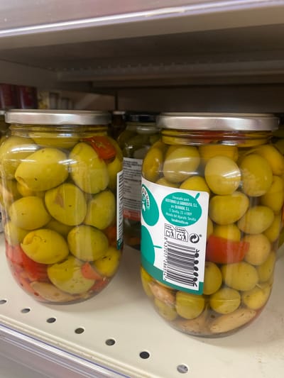 The image shows two glass jars of green olives with red bell pepper pieces, displayed on a shelf in a grocery store. The jars are clear, allowing the contents to be fully visible. The olives are plump and appear to be in brine. The top jar has a label with Spanish text and a barcode. The label reads "Envasado para ACEITUNAS LA SABROSITA, S.L. por OLIVAIDA, S.L. C/ Sevilla, 23. 41809 Albaida del Aljarafe, Sevilla lasabrosita.es" and "DE OLIVA". Below this, a barcode is visible along with the number "8 437017 286554 >". The scene is brightly lit, suggesting an indoor retail environment. The setting appears to be a supermarket aisle, with shelves and other products potentially out of frame. The focus is on the jars of olives, highlighting their contents and packaging. No people are visible in the image. The overall impression is a product shot of olives for sale.
