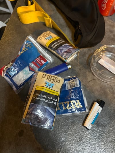 The image depicts a collection of smoking-related items scattered on a textured, dark gray surface, possibly a table or countertop, which appears to be outdoors. In the background, a yellow strap is visible, and a dark fabric bag is partially seen. A red cylindrical object, possibly a container, is also in the background.
The foreground is dominated by several packets of rolling papers and filters. One packet, prominently displayed, is yellow with "PUEBLO" written in large letters. It also shows text in Italian, "Smetti di fumare - Vivi per i tuoi cari" (Stop smoking - Live for your loved ones), along with a health warning image of a person. Another blue packet, also with "PUEBLO" branding, is labeled "WORLD'S NO.1 ROLLING PAPERS". A packet of "SLIM X-LONG" filters, with "100 FILTERS" and a diameter of "6 MM", is also visible, as is a packet of "BROWN" rolling papers.
A blue lighter lies next to the packets, and a clear glass ashtray with a single white rolling paper inside is situated to the right. A white and black disposable lighter is also present in the lower right corner. The overall scene suggests a casual setting where these items are laid out, potentially before or after use.
The specific location in the city of Ospedaletti, Italy, cannot be determined from the image alone.