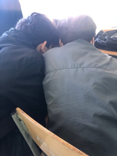 The image displays two individuals from a rear, slightly elevated perspective, situated indoors in Ortabatumi, Georgia. The person on the left has dark hair, wears a dark jacket, and is leaning with their head turned towards the right, revealing part of their ear. The person on the right, with lighter brown hair, wears a grey or sage green jacket featuring a prominent horizontal seam across the upper back, and is leaning with their head turned towards the left. Their heads are in close proximity, indicating a direct interaction or shared focus. A strong light source from the upper background creates significant backlighting and glare, obscuring details beyond the immediate subjects. In the lower foreground, a light-colored wooden desk or chair back, fitted with a visible metal support mechanism, partially obstructs the view. A dark bag, likely a backpack, is visible on a surface in the upper right background, consistent with an educational or study environment.